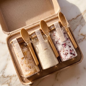 Könnte beinhalten: Ein Set aus drei Glasbehältern mit Badesalz, jeweils mit einem Holzdeckel und einem kleinen Holzlöffel, der mit einem transparenten Band befestigt ist. Die Gläser befinden sich in einem Karton und sind mit Stress & Anxiety Relief Soak, Soothing Oat & Lavender und Coconut Rose Beauty Bath Soak beschriftet, inklusive Anweisungen zur Anwendung im warmen Badewasser.
