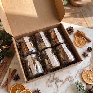 May include: A cardboard gift box containing six small glass jars filled with dried botanicals, orange slices, and spices. Each jar features a white paper label with text. The set is nestled in shredded paper filler, surrounded by dried orange slices and cinnamon sticks.