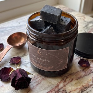 May include: An amber glass jar filled with dark grey, square salt exfoliating cubes. The label reads Moonshadow, Salt Exfoliating Cubes, Bergamot, Rock Rose, Musk. A copper-colored measuring spoon and dried dark red rose petals are nearby.