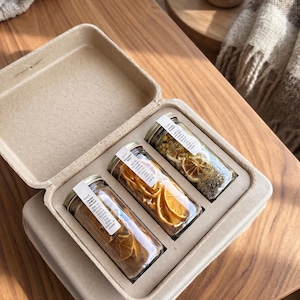 May include: A light brown molded fiber tray containing three clear glass jars with metal lids. Each jar is filled with dried botanical ingredients, including sliced citrus, herbs, and flowers, and features a white rectangular label with text.