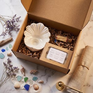 May include: A gift box containing a white ceramic shell-shaped candle holder, a small glass vial of matches, and a rectangular box of six wax melts labeled with scents including Sirens, Salt & Sea, and Black Sea, nestled in brown paper filler.