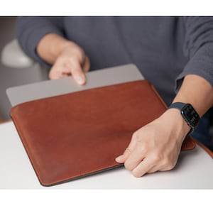 Pode incluir: Uma capa para laptop de couro marrom com um laptop cinza dentro. A capa tem uma borda costurada e está sendo segurada pela mão de uma pessoa.