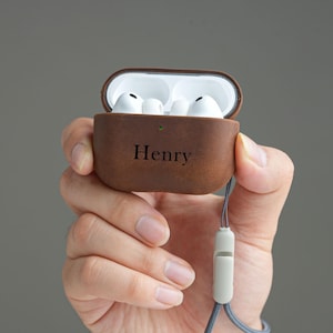 May include: A brown leather protective case for wireless earbuds with the name Henry embossed in black on the front. The case is open, revealing white earbuds inside, and features a small green indicator light and a grey lanyard attachment.