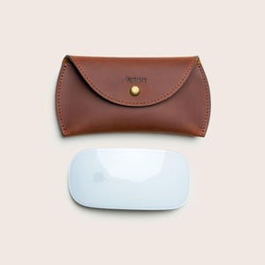 May include: A brown leather case with the name "Jason" embossed on the front, secured by a gold-colored snap. Below it is a white, rectangular computer mouse with a silver apple logo.