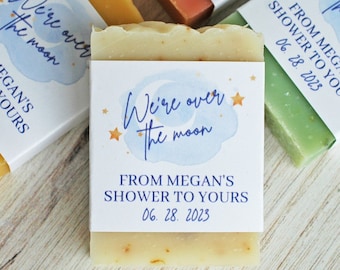 May include: Rectangular bars of soap in various colors, each wrapped in a white paper band featuring a blue watercolor cloud, orange stars, and the text: We're over the moon, FROM MEGAN'S SHOWER TO YOURS, 06. 28. 2023, printed in dark blue ink.