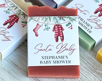 Op de afbeelding: Rechthoekige stukken zeep verpakt in witte papieren wikkels met een aquarelillustratie van een waslijn met een rood gestreept rompertje, kousen en dennentakken. De tekst luidt Santa Baby en Stephanie's Baby Shower in donker schrift.