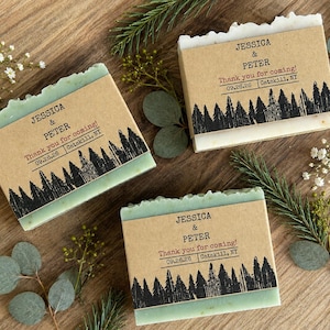 May include: Three rectangular bars of soap wrapped in tan paper bands. Each band features a black forest silhouette print and the text: JESSICA & PETER, Thank you for coming!, 09.26.26, Catskill, NY. The soap bars have a light green hue.