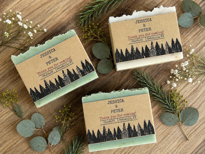 May include: Three rectangular bars of soap wrapped in tan paper bands. Each band features a black forest silhouette print and the text: JESSICA & PETER, Thank you for coming!, 09.26.26, Catskill, NY. The soap bars have a light green hue.