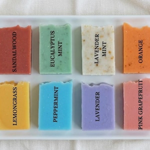 Puede incluir: Una bandeja rectangular blanca contiene ocho jabones artesanales de colores. Cada barra est&aacute; etiquetada con un aroma: S&aacute;ndalo, Menta Eucalipto, Menta Lavanda, Naranja, Hierba Lim&oacute;n, Menta Piperita, Lavanda y Pomelo Rosa. Los jabones son de varios colores.