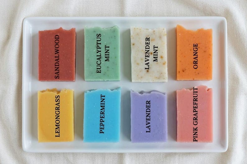 Puede incluir: Una bandeja rectangular blanca contiene ocho jabones artesanales de colores. Cada barra est&aacute; etiquetada con un aroma: S&aacute;ndalo, Menta Eucalipto, Menta Lavanda, Naranja, Hierba Lim&oacute;n, Menta Piperita, Lavanda y Pomelo Rosa. Los jabones son de varios colores.