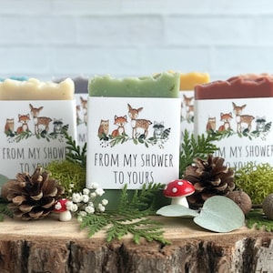 Puede incluir: Una colecci&oacute;n de jabones artesanales de varios colores, cada uno envuelto con una etiqueta con animales del bosque y el texto "FROM MY SHOWER TO YOURS". Los jabones se exhiben sobre una superficie de madera con pi&ntilde;as, vegetaci&oacute;n y un peque&ntilde;o hongo.