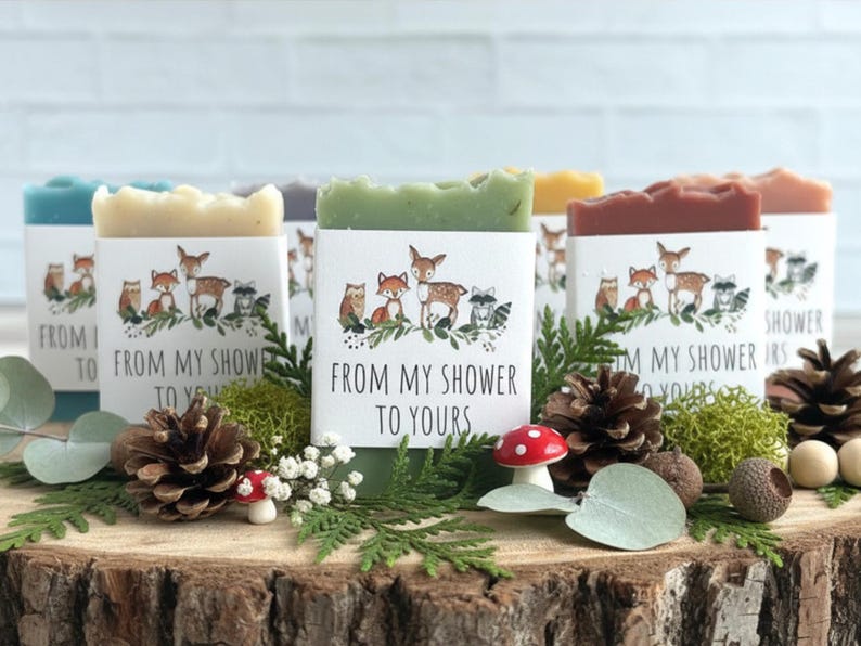 Puede incluir: Una colecci&oacute;n de jabones artesanales de varios colores, cada uno envuelto con una etiqueta con animales del bosque y el texto "FROM MY SHOWER TO YOURS". Los jabones se exhiben sobre una superficie de madera con pi&ntilde;as, vegetaci&oacute;n y un peque&ntilde;o hongo.