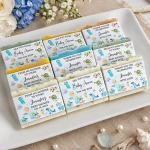 May include: A collection of rectangular soap bars arranged on a white rectangular tray. Each soap is wrapped in a paper label featuring colorful watercolor illustrations of sea creatures and text including Sea You Soon Baby Aaron or Jennifer.