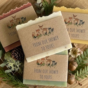 Puede incluir: Cuatro pastillas de jab&oacute;n en tonos rojo, blanco, amarillo y verde, cada una envuelta con una banda de papel marr&oacute;n. Las bandas presentan una ilustraci&oacute;n de animales del bosque y el texto "FROM OUR SHOWER TO YOURS". Los jabones est&aacute;n dispuestos sobre una superficie de madera con vegetaci&oacute;n.