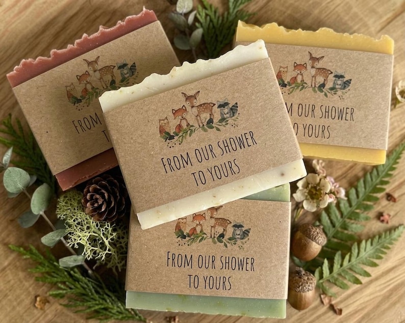 Puede incluir: Cuatro pastillas de jab&oacute;n en tonos rojo, blanco, amarillo y verde, cada una envuelta con una banda de papel marr&oacute;n. Las bandas presentan una ilustraci&oacute;n de animales del bosque y el texto "FROM OUR SHOWER TO YOURS". Los jabones est&aacute;n dispuestos sobre una superficie de madera con vegetaci&oacute;n.