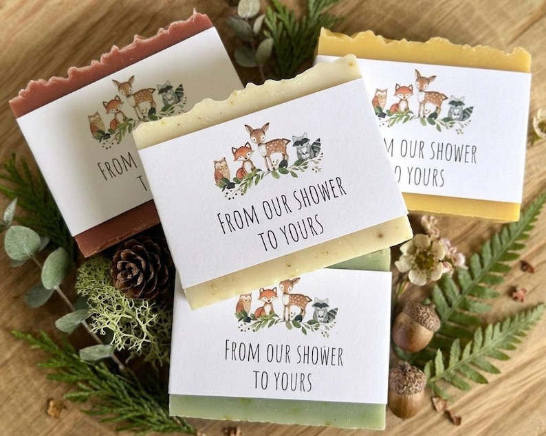 Puede incluir: Cuatro barras de jab&oacute;n hechas a mano con etiquetas que dicen "FROM OUR SHOWER TO YOURS". Cada etiqueta presenta una ilustraci&oacute;n de animales del bosque. Los jabones son de varios colores, incluyendo marr&oacute;n, amarillo y verde.