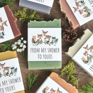 Puede incluir: Surtido de jabones artesanales de varios colores, cada uno envuelto con una banda de papel con ilustraciones de animales del bosque y el texto "FROM MY SHOWER TO YOURS". Los jabones est&aacute;n dispuestos con vegetaci&oacute;n decorativa.