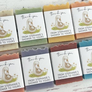 Puede incluir: Surtido de jabones artesanales de varios colores, cada uno envuelto con una etiqueta con un cisne y el texto "Thank you, From Stephanie's Shower to Yours." Los jabones est&aacute;n dispuestos sobre una superficie de madera clara.