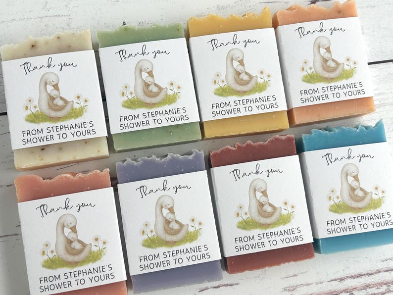 Puede incluir: Surtido de jabones artesanales de varios colores, cada uno envuelto con una etiqueta con un cisne y el texto "Thank you, From Stephanie's Shower to Yours." Los jabones est&aacute;n dispuestos sobre una superficie de madera clara.