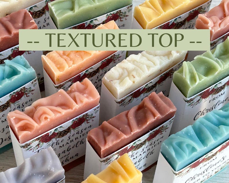 May include: A collection of rectangular bars of soap in various pastel colors including pink, blue, yellow, white, and green. Each bar features a textured top and is wrapped in a white paper band with floral graphics and printed text.