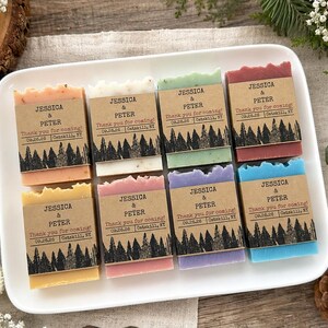 May include: Eight rectangular bars of soap in various colors, each wrapped in a brown paper band featuring a black forest silhouette graphic and the printed text: JESSICA & PETER, Thank you for coming!, 09.26.26, Catskill, NY, arranged on a white tray.