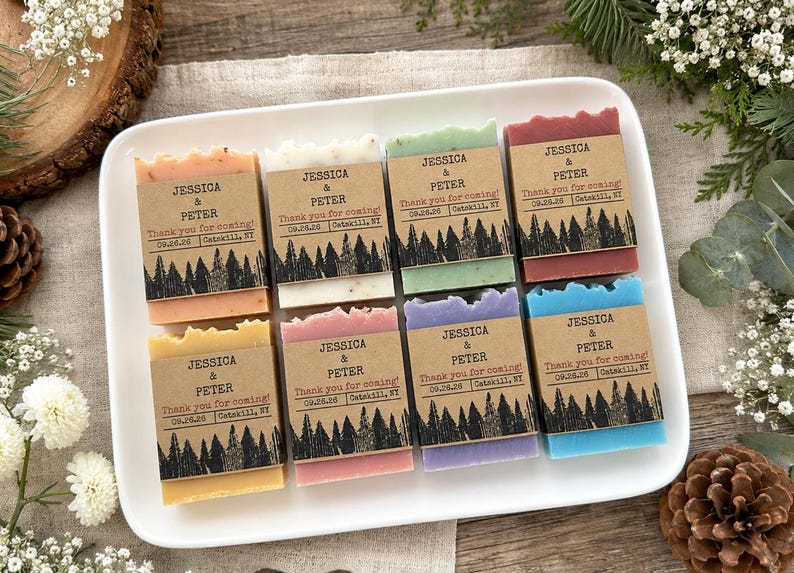 May include: Eight rectangular bars of soap in various colors, each wrapped in a brown paper band featuring a black forest silhouette graphic and the printed text: JESSICA & PETER, Thank you for coming!, 09.26.26, Catskill, NY, arranged on a white tray.