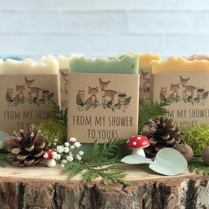 Puede incluir: Una colecci&oacute;n de jabones artesanales de varios colores, cada uno envuelto en una funda de papel kraft con un dise&ntilde;o de animales del bosque y el texto "FROM MY SHOWER TO YOURS". Los jabones est&aacute;n dispuestos sobre una rodaja de madera con pi&ntilde;as, vegetaci&oacute;n y elementos decorativos.