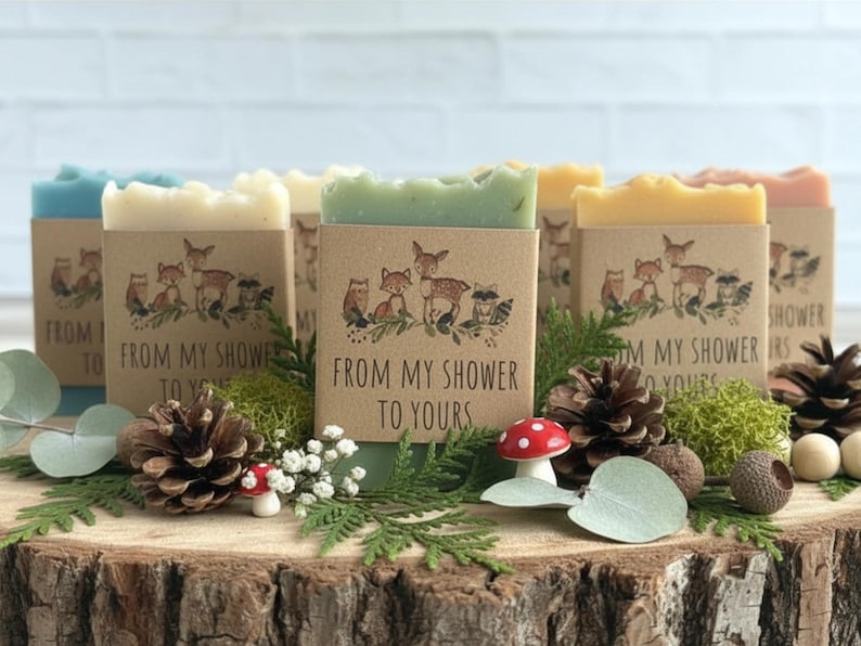 Puede incluir: Una colecci&oacute;n de jabones artesanales de varios colores, cada uno envuelto en una funda de papel kraft con un dise&ntilde;o de animales del bosque y el texto "FROM MY SHOWER TO YOURS". Los jabones est&aacute;n dispuestos sobre una rodaja de madera con pi&ntilde;as, vegetaci&oacute;n y elementos decorativos.