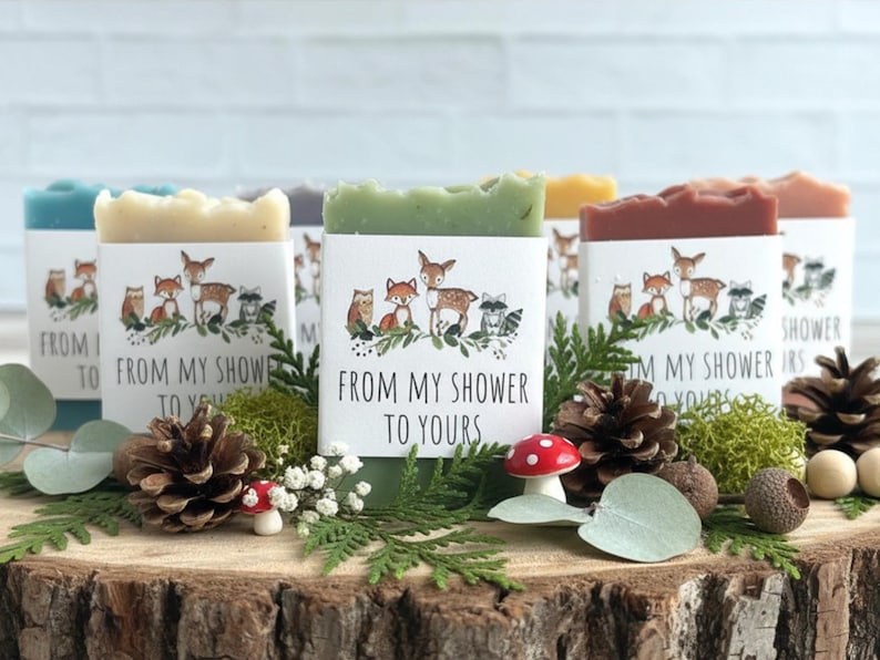 Pode incluir: Uma cole&ccedil;&atilde;o de sabonetes artesanais em v&aacute;rias cores, cada um embrulhado com um r&oacute;tulo com criaturas da floresta e o texto "FROM MY SHOWER TO YOURS". Os sabonetes s&atilde;o exibidos em uma superf&iacute;cie de madeira com pinhas, vegeta&ccedil;&atilde;o e um pequeno cogumelo.