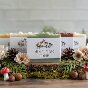 Pode incluir: Uma cole&ccedil;&atilde;o de sabonetes artesanais com r&oacute;tulos decorativos com animais da floresta. Os sabonetes s&atilde;o exibidos em uma base de madeira com detalhes florais e naturais, incluindo pinhas, cogumelos e bolotas. O r&oacute;tulo diz "FROM OUR SHOWER TO YOURS".