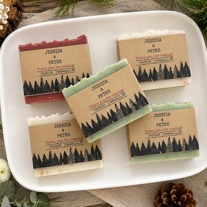 May include: Four rectangular bars of soap wrapped in brown paper bands. Each band features a black forest silhouette print and text reading Jessica & Peter, Thank you for coming!, 09.26.26, Catskill, NY. The soaps are red, green, and off-white.