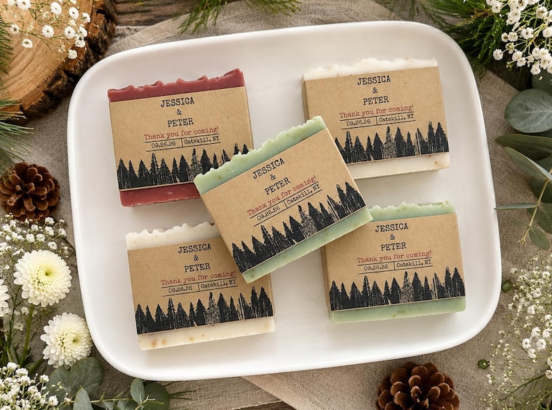 May include: Four rectangular bars of soap wrapped in brown paper bands. Each band features a black forest silhouette print and text reading Jessica & Peter, Thank you for coming!, 09.26.26, Catskill, NY. The soaps are red, green, and off-white.