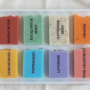 May include: Eight rectangular bars of soap arranged on a white rectangular tray. The soaps are labeled Sandalwood, Eucalyptus Mint, Lavender Mint, Orange, Lemongrass, Peppermint, Lavender, and Pink Grapefruit in black text on each bar.
