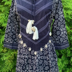 May include: A navy blue dress with white floral and geometric patterns. The dress features long sleeves with a floral design, a chevron-patterned bodice, and is adorned with white tassels and seashells.