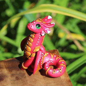 Peut inclure: Une petite figurine de dragon rose avec des accents blancs et dorés. Le dragon a une queue enroulée et est assis sur une surface en bois.