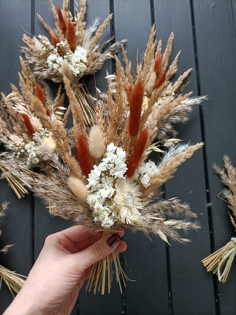 Pu&ograve; includere: Bucaneve di fiori secchi con un mix di erbe e fiori beige, marroni e color ruggine. I bouquet sono legati con spago naturale e includono accenti bianchi. L'immagine mostra diversi bouquet, uno dei quali &egrave; tenuto in mano.