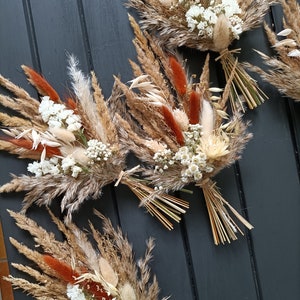 Pu&ograve; includere: Diversi bouquet di fiori secchi con un mix di erbe e fiori beige, marroni e color ruggine. Ogni bouquet &egrave; legato con spago naturale e presenta piccoli fiori bianchi. I bouquet sono disposti su una superficie grigio scuro.