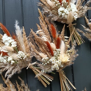 Pu&ograve; includere: Primo piano di diversi bouquet di fiori secchi. I bouquet presentano un mix di erbe beige, marroni e arancioni, accentate da piccoli fiori bianchi. Ogni bouquet &egrave; legato con spago e paglia.