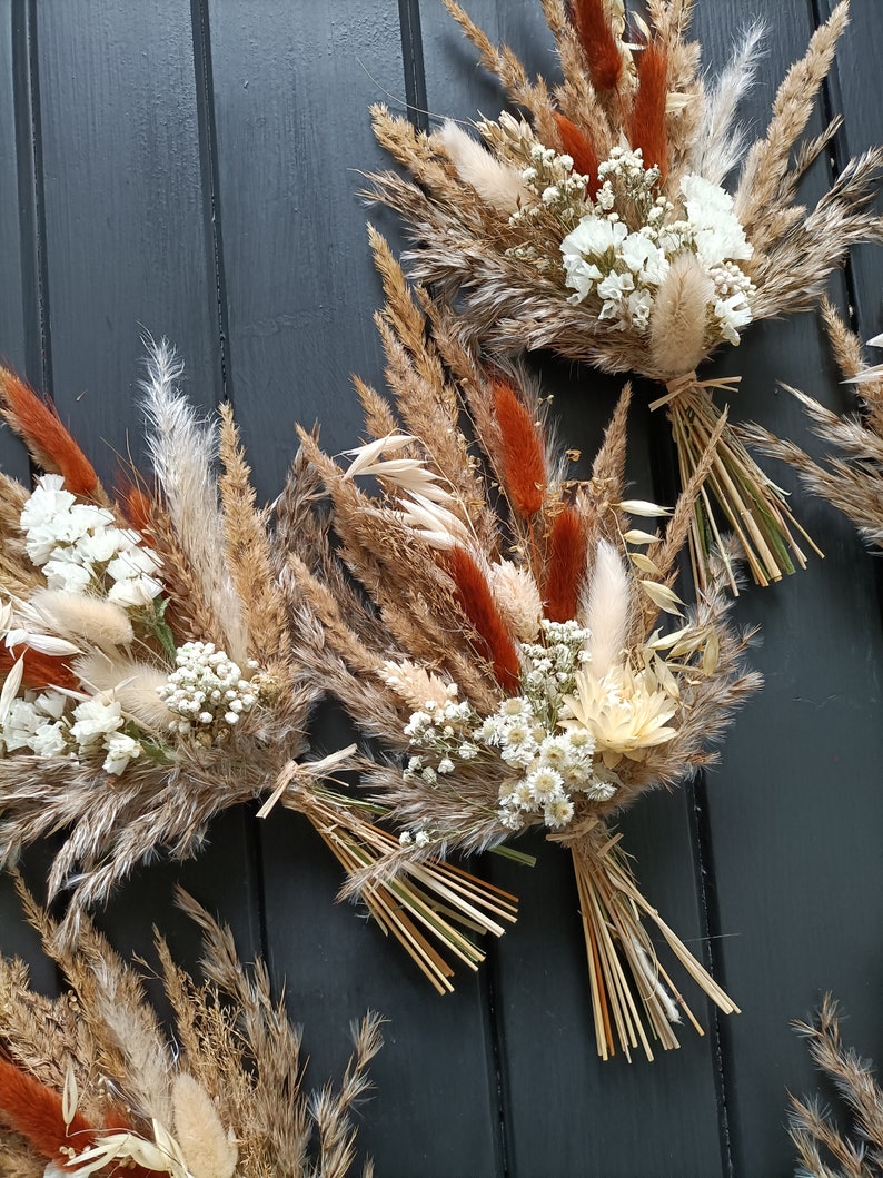 Pu&ograve; includere: Primo piano di diversi bouquet di fiori secchi. I bouquet presentano un mix di erbe beige, marroni e arancioni, accentate da piccoli fiori bianchi. Ogni bouquet &egrave; legato con spago e paglia.