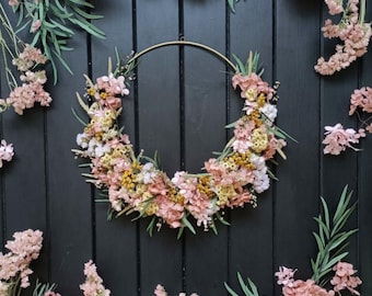 May include: A floral wreath with a gold metal ring base, adorned with pink, yellow, and white dried flowers and greenery. The wreath is set against a black wooden background, with additional floral arrangements scattered around it.