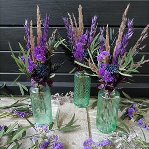 Peut inclure: Trois vases en verre texturés, remplis de fleurs séchées violettes et de verdure. Les vases sont disposés sur une surface en bois.