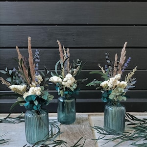 Puede incluir: Tres jarrones de cristal color turquesa con diseños texturizados, cada uno con un pequeño arreglo de flores secas y vegetación. Los arreglos incluyen elementos blancos, lavanda y beige. Los jarrones se exhiben sobre una superficie clara.