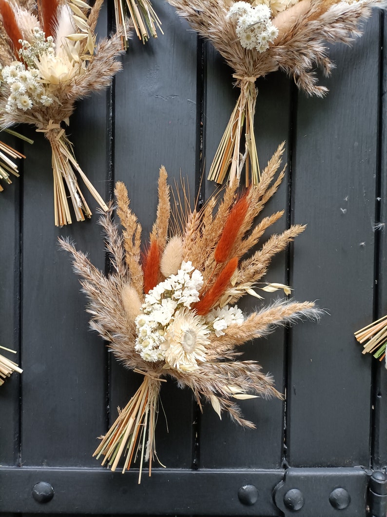 Pu&ograve; includere: Primo piano di bouquet di fiori secchi. I bouquet sono composti da grano, erba della pampa e altri fiori secchi in tonalit&agrave; beige, marrone e arancione. Legati con spago naturale, questi bouquet sono un elemento decorativo rustico.