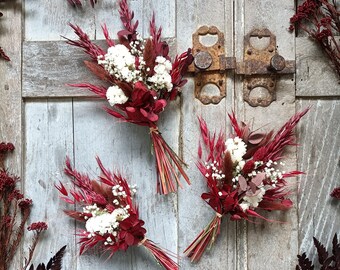 Può includere: Tre piccoli mazzi di fiori secchi in tonalità di rosso e bianco. I mazzi sono disposti su una superficie di legno rustica.