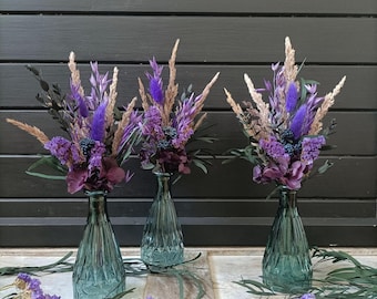 Può includere: Tre vasi in vetro con motivo a diamante color verde acqua, ciascuno contenente una composizione di fiori secchi. I bouquet presentano piume viola, bacche blu scuro, petali di ortensia viola e steli di grano beige.