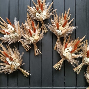 Pu&ograve; includere: Diversi piccoli bouquet di fiori secchi. Ogni bouquet presenta un mix di erbe e fiori beige, marroni e color ruggine, legati con paglia. I bouquet sono disposti su uno sfondo di legno grigio scuro.