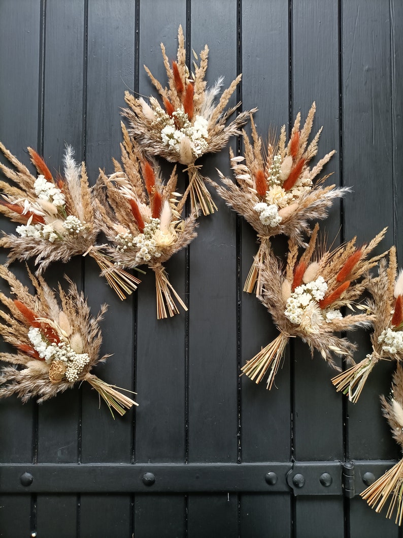 Pu&ograve; includere: Diversi piccoli bouquet di fiori secchi. Ogni bouquet presenta un mix di erbe e fiori beige, marroni e color ruggine, legati con paglia. I bouquet sono disposti su uno sfondo di legno grigio scuro.