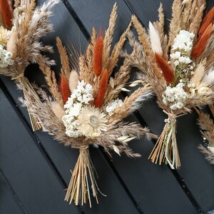Pu&ograve; includere: Primo piano di diversi bouquet di fiori secchi con grano, erba della pampa e fiori bianchi. I bouquet sono legati con spago e hanno accenti arancioni e beige.