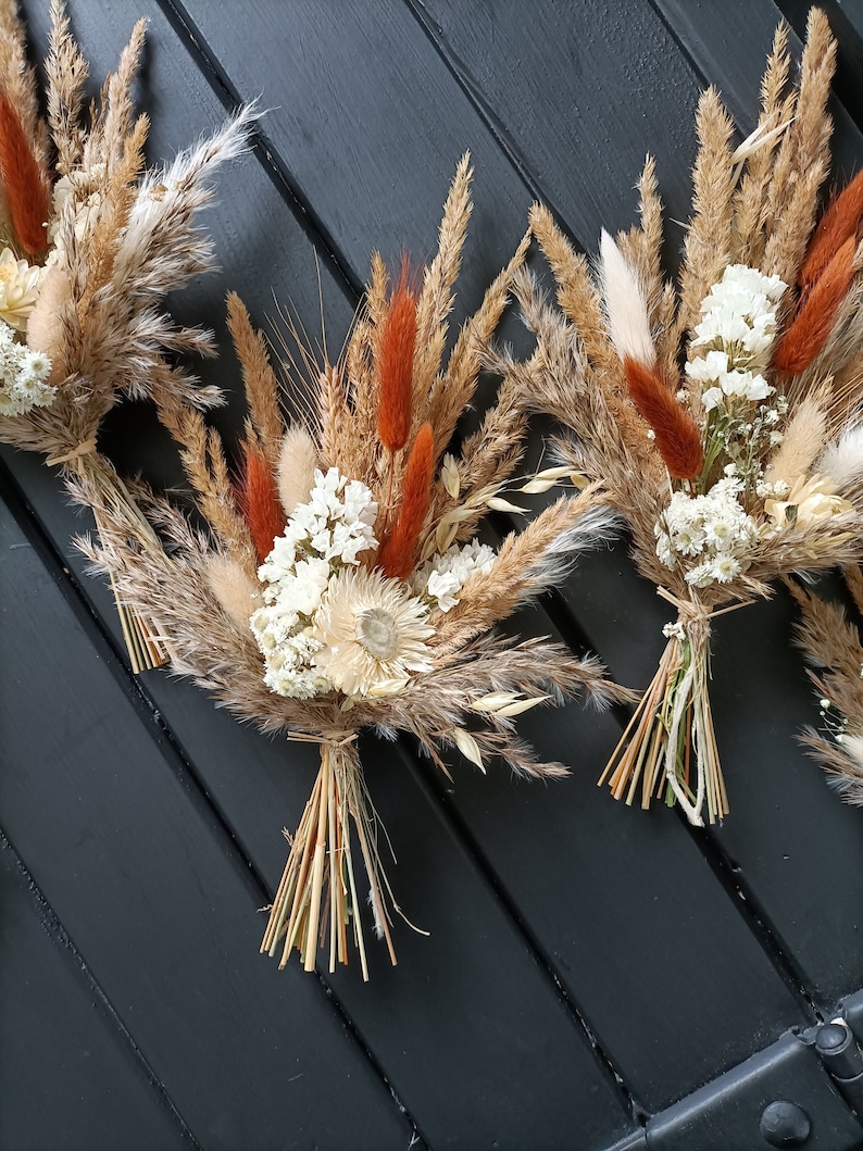 Pu&ograve; includere: Primo piano di diversi bouquet di fiori secchi con grano, erba della pampa e fiori bianchi. I bouquet sono legati con spago e hanno accenti arancioni e beige.