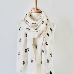 Peut inclure: Une &eacute;charpe blanche avec des motifs en forme de feuilles noires et brunes et des glands. L'&eacute;charpe est drap&eacute; sur un mannequin blanc.